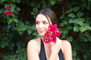 Young beautiful brunette typical Spanish woman dressed in black suit and bouquet of pink flowers near her face. The woman is in the park surrounded by greenery. The woman is happy.