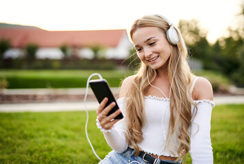 Young happy teenager girl sitting in the grass and texting on a smart phone while listening to music on headphones - Mobile technology and modern entertainment concept