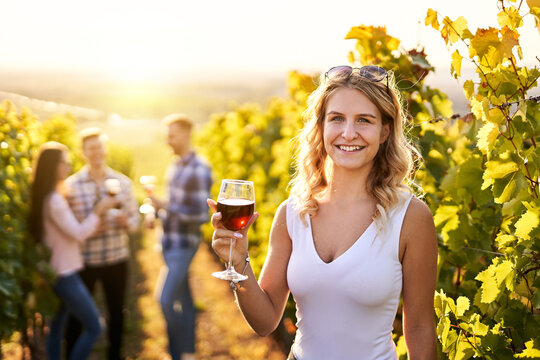 Portrait of a young, millennial woman holding a glass of organic bio red wine outdoors in a vineyard with her friends in the background - Vine-growing, and wine-tasting concept
