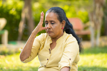 Indian senior woman doing nostril breathing exercise or pranayama yoga with eyes closed at park -...