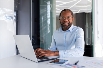Fototapeta premium Portrait of mature businessman inside modern office, senior man in shirt smiling and looking at camera, worker using laptop at work