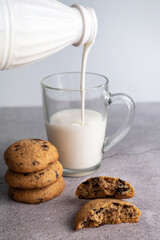 On a gray table are cookies with chocolate, a glass cup into which milk is poured from a bottle.