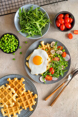 Fried egg with waffles on a plate with salad - arugula, tomatoes and radish with mushrooms. Top view, ingredients in bowls on the table.