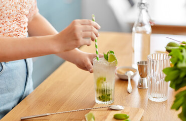 drinks and people concept - close up of woman with paper straw making lime mojito cocktail at home...