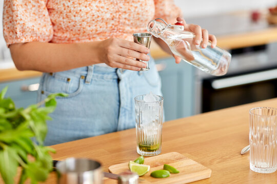 drinks and people concept - close up of woman pouring water from glass bottle to jigger and making lime mojito cocktail at home kitchen