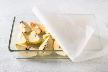 Quartered quince in a glass baking dish covered with baking paper on a light gray background. Stage of preparation of vegan quince pastila or other dessert