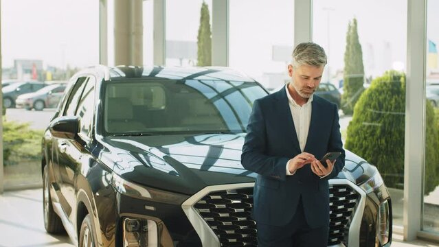Portrait of a middle age mature handsome beard grey haired man in a suit using smartphone leaning on car and thinking if he should buy it. Businessman standing next to new car.