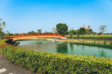 Fototapeta premium Beautiful view of Japanese style red wooden bridge over emerald pond with fountains against nature of paddy field outdoors. Japanese style garden with unique red wooden bridge. Beautiful landscape.