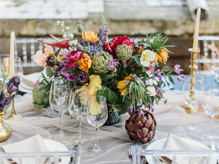 Decor wedding banquet table in tropical style. A colorful bouquet of artichoke, white and pink peonies, tulips, iris. In the foreground are wine glasses, candlesticks with candles, crockery.