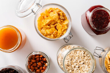 food storage and eating concept - close up of jars with oat, corn flakes, granola, cookies and spreads on white background