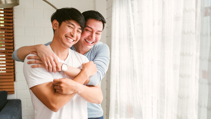 Portraits of happy millennial Asian gay couple hugging and embracing, smiling and laughing in a white curtain window in the living room at LGBT multi-relationship home. An enjoy gay couple concept.