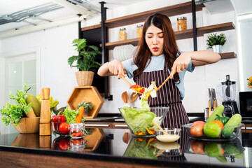 Happy Asian woman wearing apron, tossing the vegetarian salad with wooden spatulas. Preparing a healthy salad with fresh vegetables such as carrot, tomato cabbage and green oak in the home kitchen.