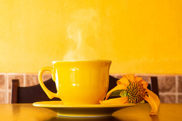 Uma xícara amarela cheia de café quente, com pires e um girassol murcho, sobre uma mesa de madeira com uma parede amarela ao fundo.