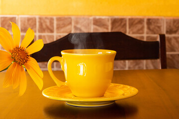 Uma xícara amarela com café quente, saindo vapor, com um girassol, sobre uma mesa de madeira.