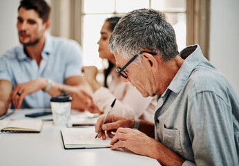Record everything, you might need it later. a mature businessman writing notes during a meeting with colleagues in a modern office.