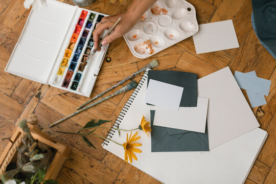 Close-up of artist sitting on the floor and painting with watercolour paints. Art supplies and blank paper sheets mockup on the floor. 