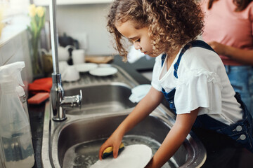 Moms got me doing the dishes. an adorable young girl standing and washing the dishes in the kitchen sink at home.