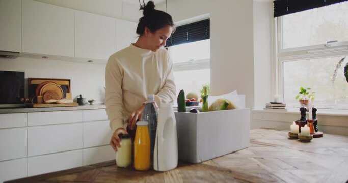 Smiling young woman unpacking a delivery box full of groceries while standing at her kitchen table