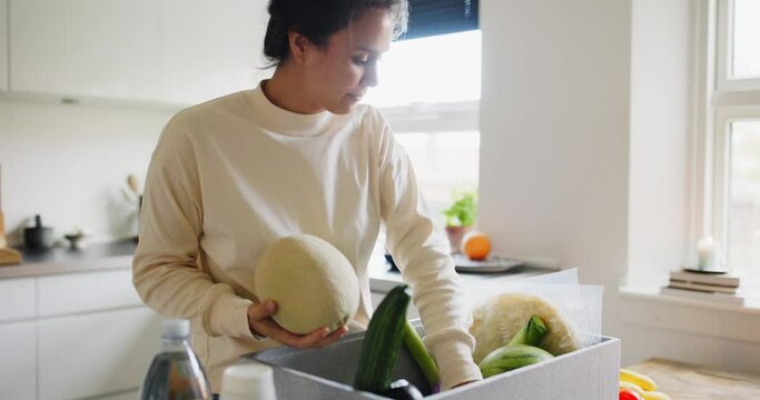 Young woman unpacking a delivery box full of groceries while standing at her kitchen table