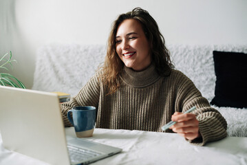 Successful female CEO multitasking at kitchen table during video conference with partners, using laptop, phone, and coffee cup