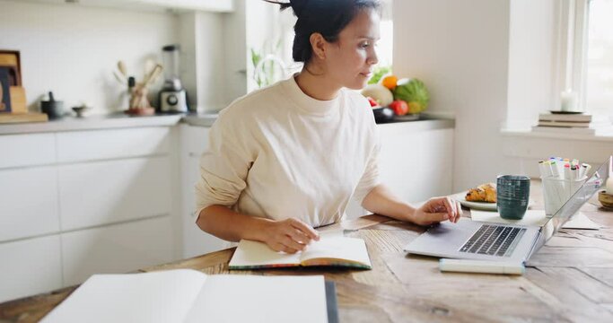 Young woman drawing in a sketchbook and working on a laptop while sitting at a table in her kitchen at home