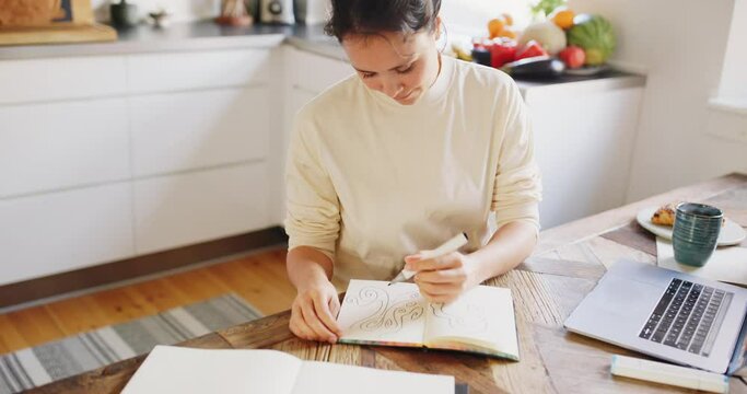 Smiling young woman drawing designs in a sketchbook while sitting at a table in her kitchen at home