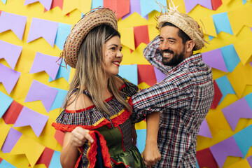 Fototapeta na wymiar Brazilian couple wearing traditional clothes for Festa Junina - June festival - dancing over colorful background with pennants