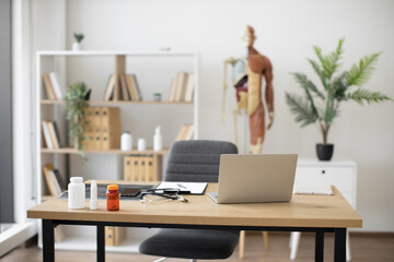 Fototapeta na wymiar Focus on writing desk with modern laptop, stethoscope, and medicine bottles against background of human skeleton model at doctor's office. Spacious consulting room in outpatient department.