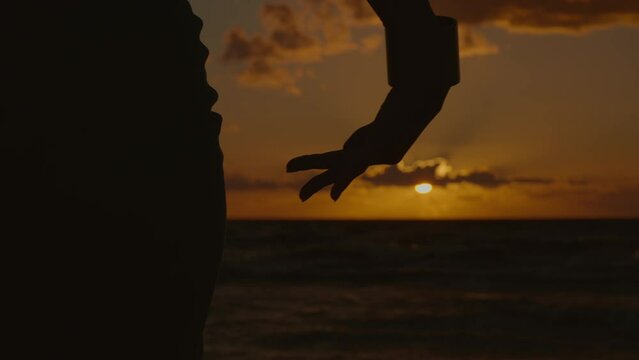 hand held close up shot of a silhouette of a woman's hand dancing beautifully at sunset by the sea, 4k, slow motion