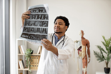 Mindful multicultural person in lab coat holding CT image of brain scan while standing in professional office of outpatient clinic. Thoughtful expert examining diagnostic test results in workplace.