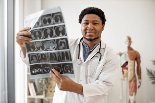 Confident african american man in doctor's coat with stethoscope holding MRI scans of human brain in consulting room on skeleton model background. Professional physician analysing diagnostic report.