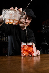 Bartender pours mixed traditional Negroni cocktail in the glass at the bar. Bartender mixes gin, campari, and sweet vermouth to prepare the Negroni alcoholic cocktail.