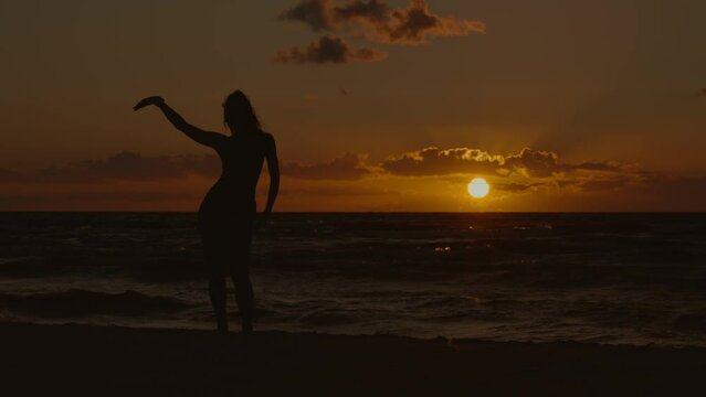 static wide shot of a woman in silhouette dancing by the sea against the background of the setting sun, 4k, slow motion