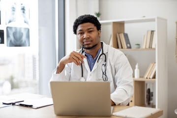 Pensive multiethnic male therapist in white coat posing with pen in hand behind computer on writing...