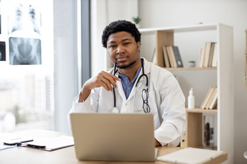 Pensive multiethnic male therapist in white coat posing with pen in hand behind computer on writing desk in doctor's office interior. Healthcare practitioner exploring diseases using technologies.