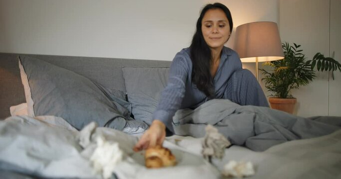 Young woman in pajamas writing and eating a snack while lying on her bed full of tissues with a cold