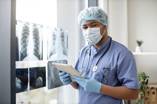 Focused multiethnic male worker in face mask and scrub top posing with digital tablet near X-ray scans fixed on window glass. Qualified surgeon in medical gloves studying injuries scrubs.