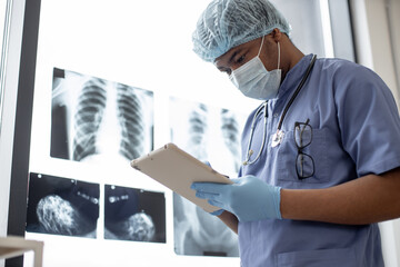 Confident multicultural doctor in scrubs and face mask using tablet while standing in hospital's office with radiography images. Male physician in protective gloves and cap sharing data with staff.
