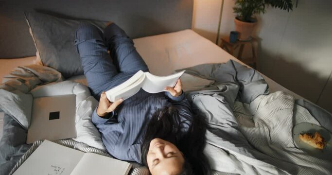 Smiling young woman wearing pajamas relaxing on her bed in the evening and reading a book