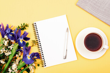 Coffee cup, blank notepad with pen and bouquet of blue iris flowers on yellow table. Top view, flat lay, mockup