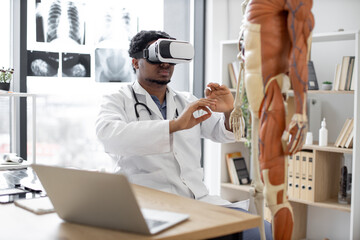 African american man wearing lab coat and 3d goggles using VR system while sitting near modern devices and human skeleton in office. Therapist preparing for medical training in teaching hospital.