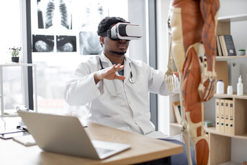 Fototapeta na wymiar African american man wearing lab coat and 3d goggles using VR system while sitting near modern devices and human skeleton in office. Therapist preparing for medical training in teaching hospital.