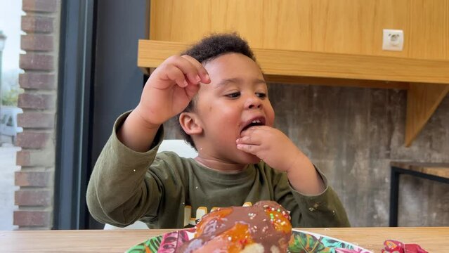 Funny and adorable 3 year-old child eating a cake covered with chocolate in a cafeteria seated next to his mother