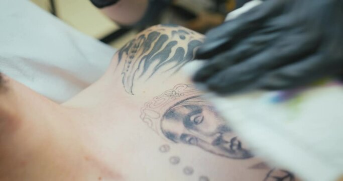 A female tattoo artist with black gloves tattoos the Virgin of Montserrat, also known as La Moreneta, on the chest of a young man with black ink using the dotwork technique. Close-up view