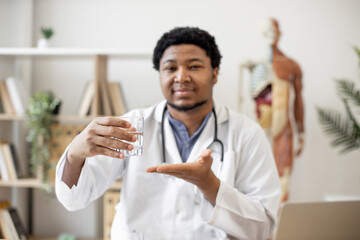 Positive multicultural physician in lab coat showing glass of water while sitting at writing desk...