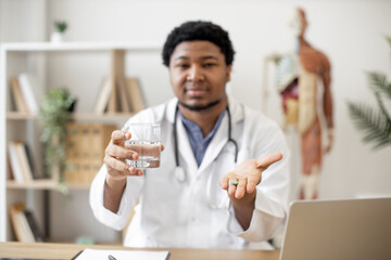 Focus on green round pill being held by multiracial male doctor sitting at writing desk with laptop on office background. Professional medical worker giving medicine and glass of water for treatment.