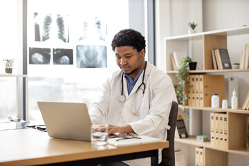 Mindful multicultural adult in white coat with stethoscope using portable computer in doctor's workplace. Health professional developing treatment plans at writing desk in teaching hospital.