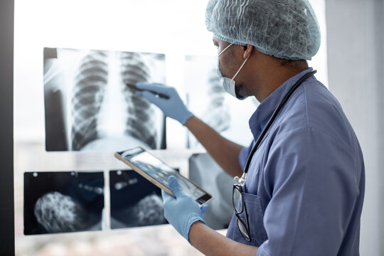 Focus on chest X-ray examination studied by multiracial healthcare practitioner in protective garment using tablet with stylus pen. Male radiologist identifying mass in left lung using technologies.