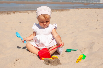 Cute blond caucasian baby girl in white dress plays with sand toys on a beach