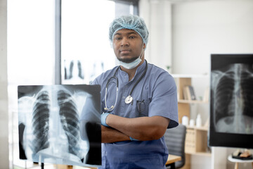 Serious multiethnic man in protective scrubs and cap posing with his arms crossed in office...
